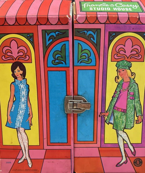 Brunette Casey in Iced Blue and blonde Francie in Tweed Somes stand on a red-and-orange tiled surface before a sparsely rendered, somewhat abstracted building with art-nouveau-style fleurs-de-lis and other flourishes with a stained glass appearance. 