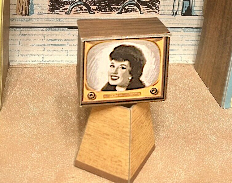 Close up on chipboard TV on stand. The set is wood-look with gold-toned front. Smiling woman on front has short bouffant hair and high fur collar that merge together in the black-and-white color scheme.