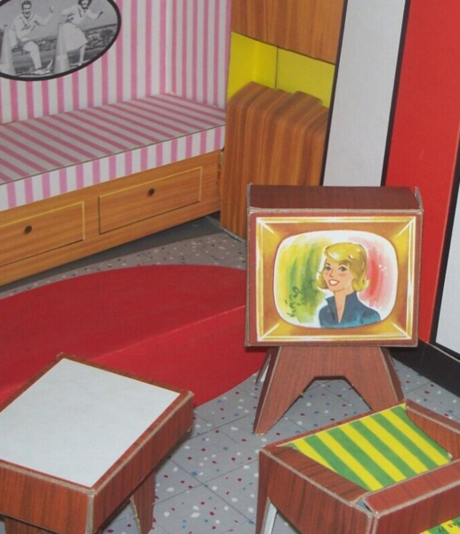 Close up on another wood-look chipboard TV with gold front, this one on four splayed legs. In color on screen is a blonde woman smiling, wearing navy with collar standing up. Behind her are washes of red and green. Surrounding we can see a dotted tile floor, a pink bench along the wall with drawers beneath, a wood-look chair with yellow-and-green striped upholstery, and a wood-look white-topped side table.