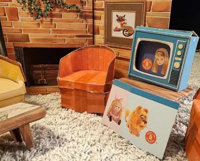 New Dream House chipboard living room. On blue "television" with insert screen, Illustrated Skipper's head floats on blue background next to Mattel logo. Alternate screens are propped against the table on which the TV sits. Both have white backgrounds; one shows a toy-looking lion and the other a little girl doll with blonde hair and pink frills. Also in frame are orange armchairs, yellow sofa, wood-look coffee table with marble-look top, and fieldstone fireplace with hanging greenery and cute framed cat picture.
