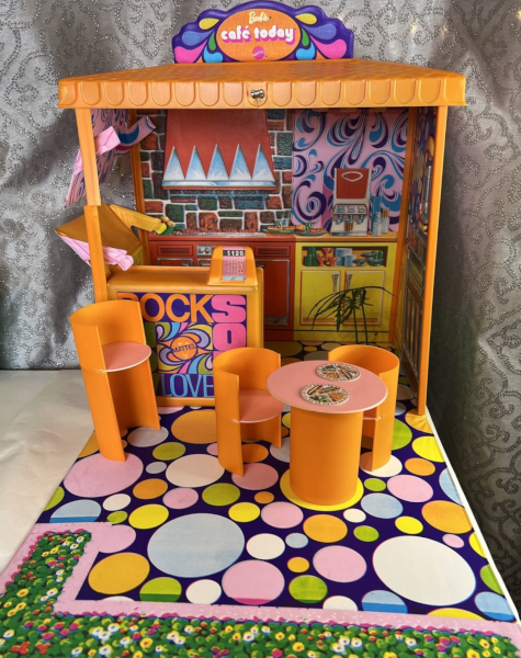 Color photo of plastic play set. Orange plastic rectangular structure has lithographed vinyl surfaces showing a grill with range hood, pink psychedelic swill wallpaper, soda fountain, and many-sized polka dot floor that can be folded up to close the box. A plastic counter is printed with the words "Now", "Rock", "Soul", and "Love", surrounding more psychedelic swirls, with cash register atop. One side of the structure has a window opening midway up, one flap folding down to form a counter and one folding up to make an awning. Three orange chairs--one at counter height, and two lower--are cylinder-shaped with flat circular seats. A circle table with thick cylindrical base has two flat plates on top printed with food (not detailed at this magnification but consisting of hot dogs, fries, olives and pickles).