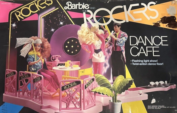 Box art from Dance Cafe shows product photo. The floor is pink and shaped like a guitar with fretboard and head. body of the "guitar" is surrounded by pink plastic fence panels with guitar shaped inset and ROCKERS written diagonally in a black stripe near the top. Two yellow directors chairs sit at a white-topped table with some dishware atop (all plastic). Along the back is a circular piece of decor resembling a record with flashing lights embedded in the center. Before that sits a pink bar. A pink plastic phone sits atop the bar, seeming a bit out of place. Starry printed elements on black background decorate the bar front and cardboard back surrounding the record decoration. A green fern in white pot sits in the foreground. Rockers Midge sits at the table, while Barbie and Ken are hooked into an embedded stand that makes them "dance" (turn) and the press of a lever.